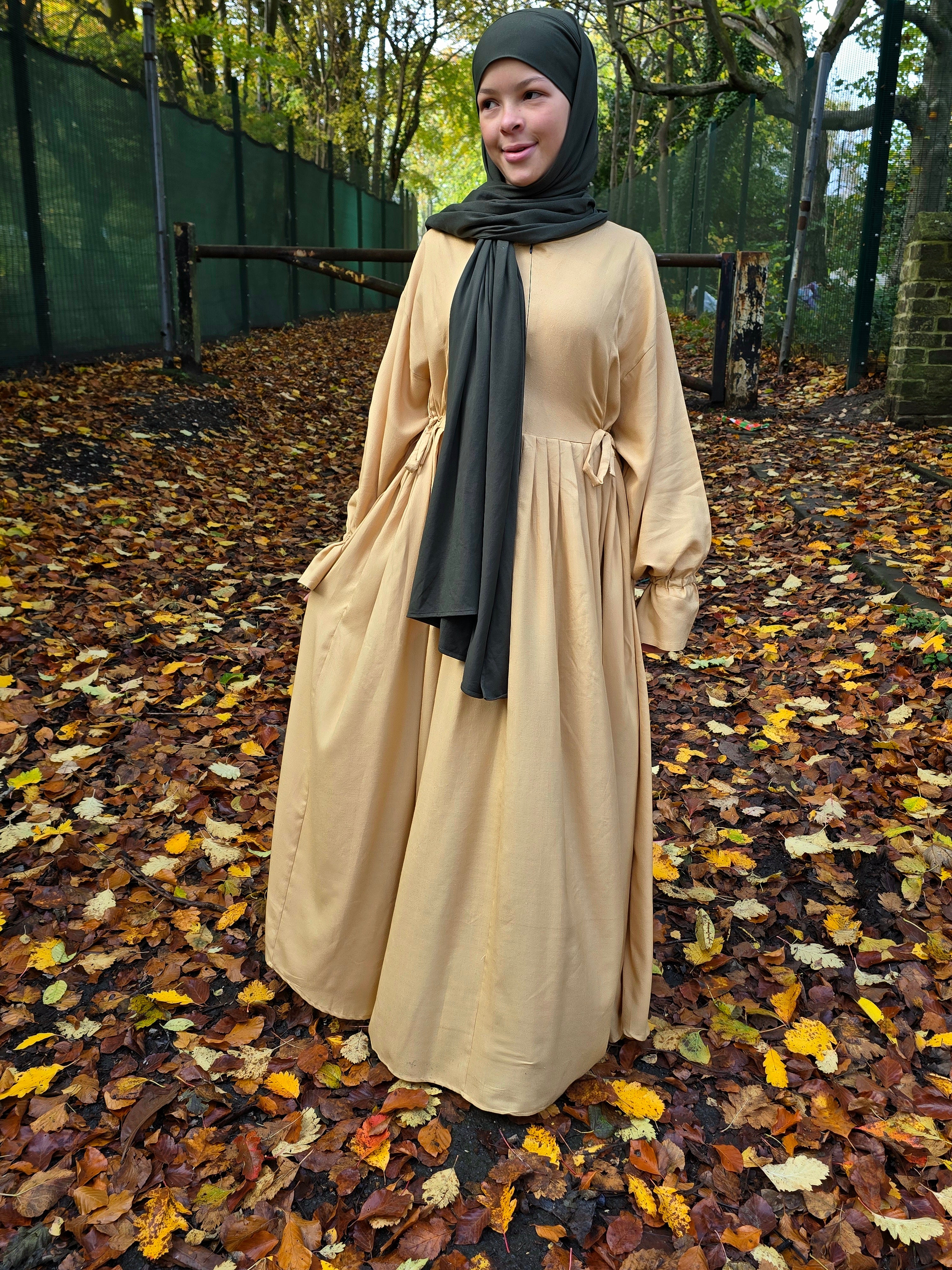 Woman in a beige abaya dress and dark green jersey hijab standing on a path with fallen leaves.