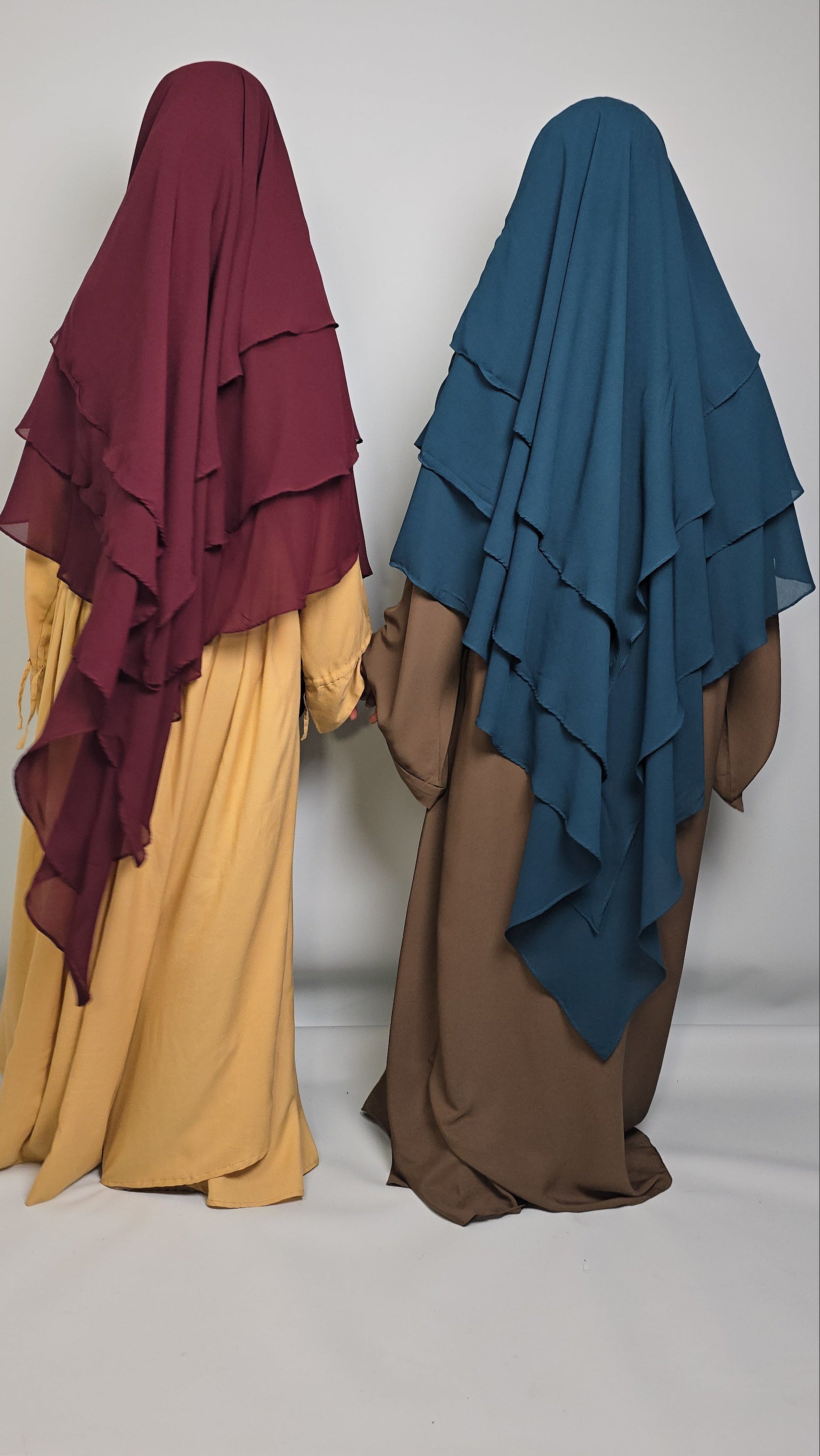 Two women one wearing a teal khimar and the other wearing a burgundy khimar on a white background