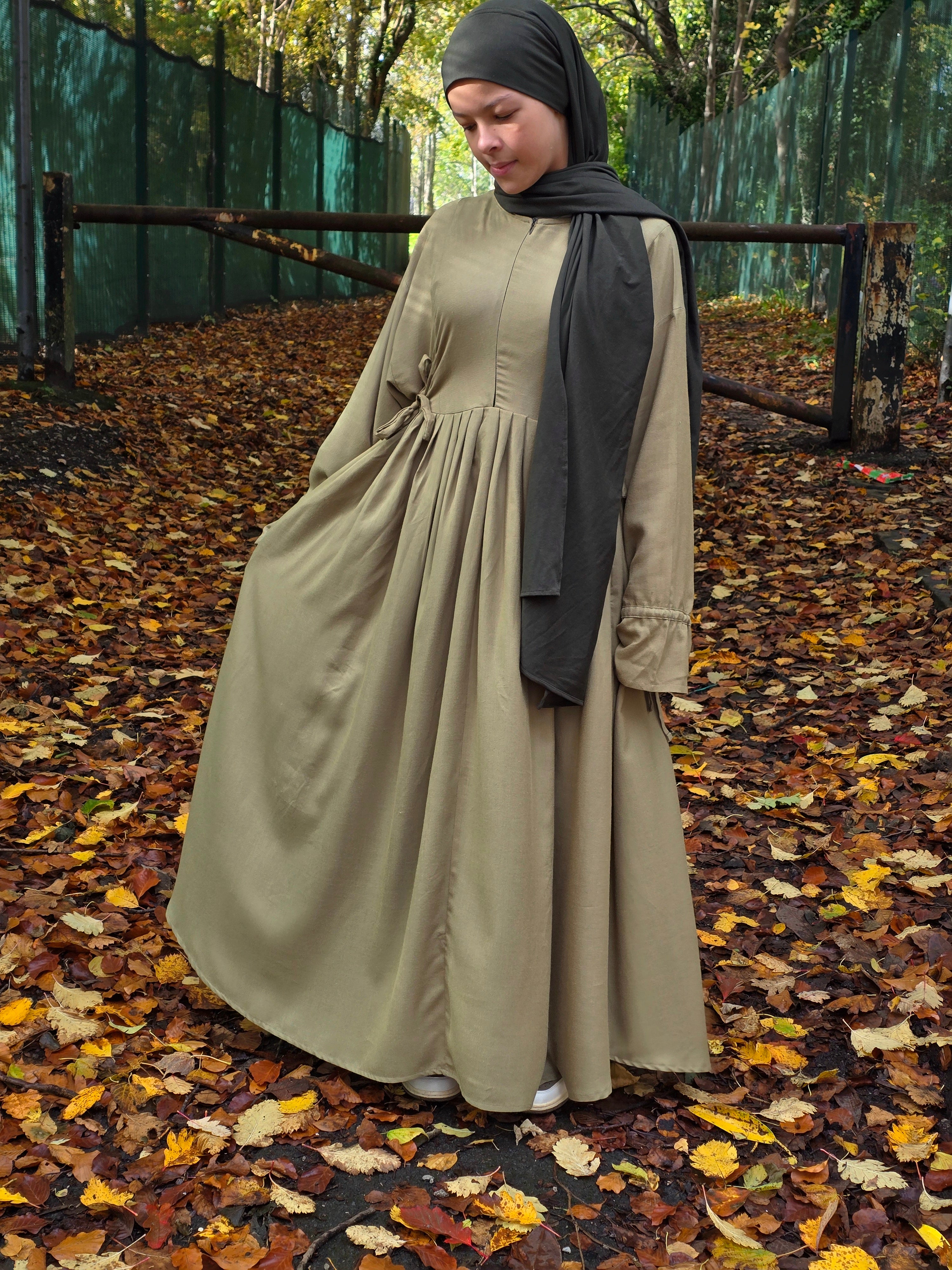 Woman in a long sage green abaya dress and headscarf standing on a path with fallen leaves.