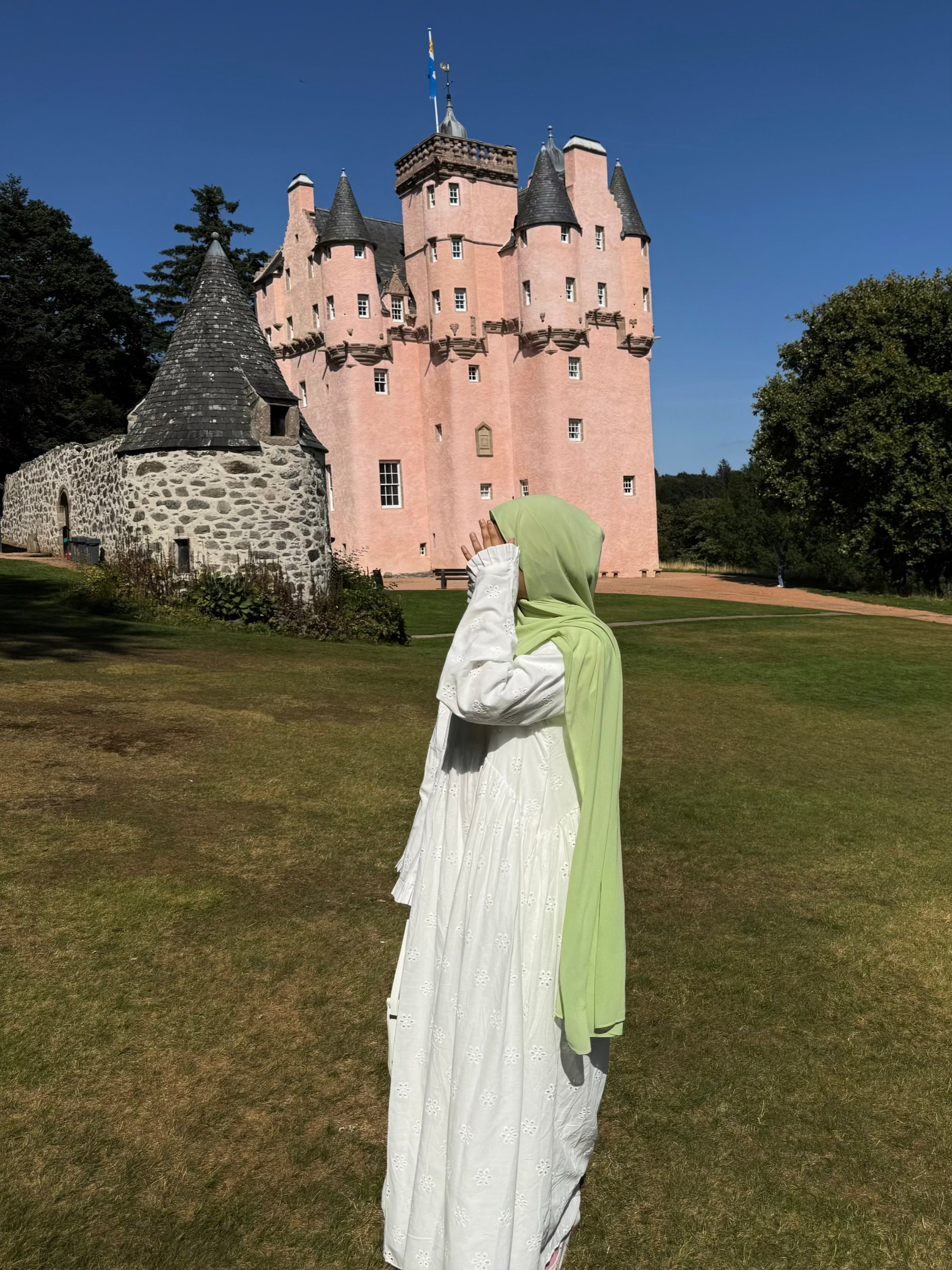 A woman in a white cotton abaya and light green hijab stands on a grassy lawn facing a large pink castle under a clear blue sky.