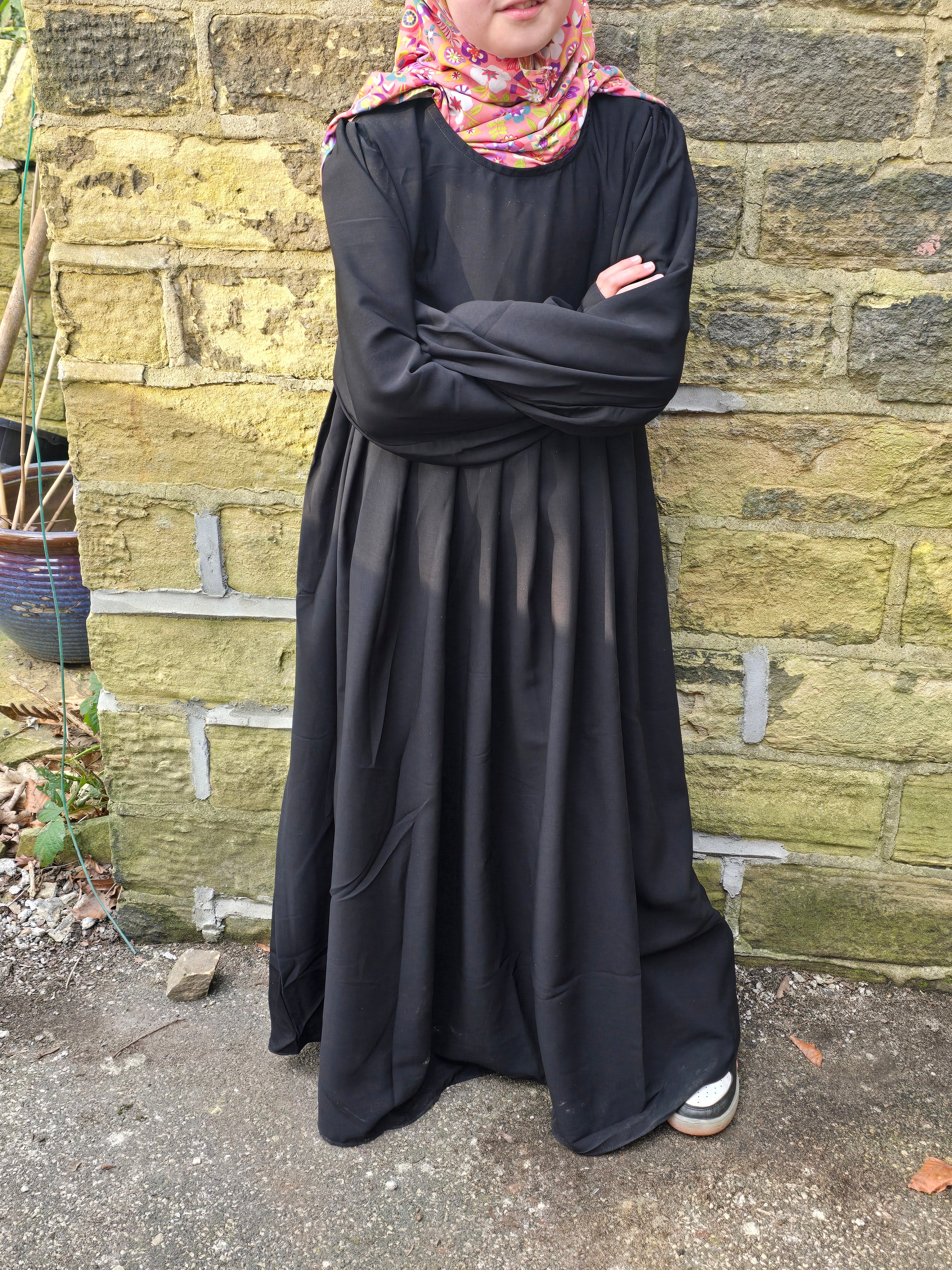 A young girl wearing a black Russian abaya with puffy sleeves and a high neckline, standing in front of a stone wall.