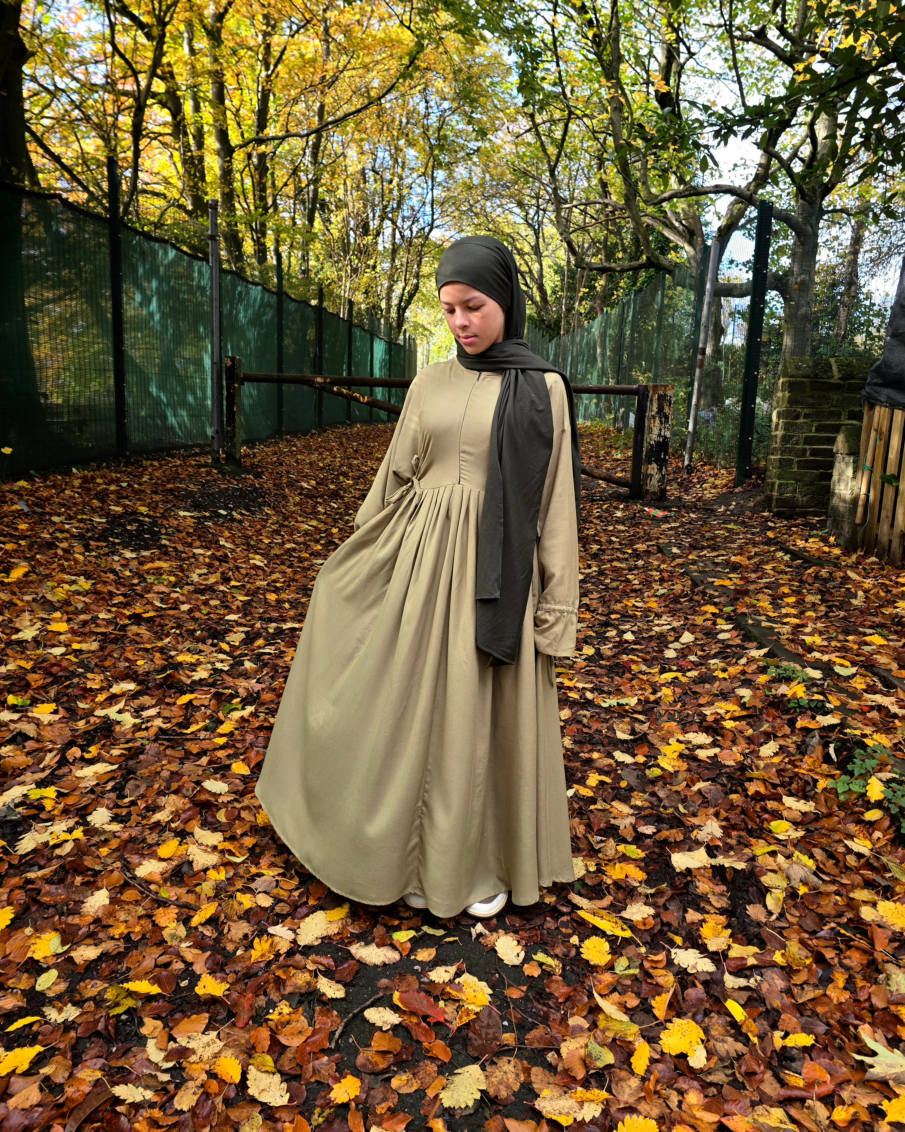 Person wearing a abaya dress and black hijab standing on a path with autumn leaves and trees.
