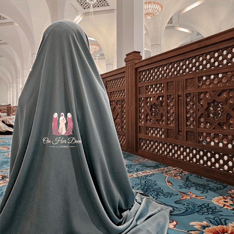 A woman wearing a pastel turquoise color two-piece jilbab with cargo pockets, standing inside a mosque.