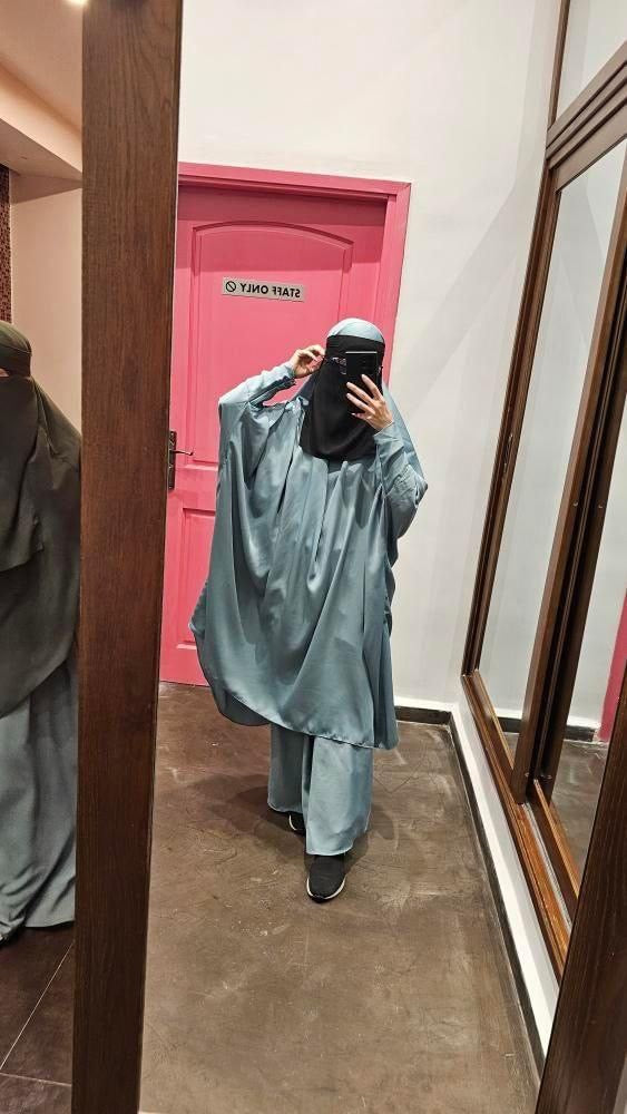 Person wearing a pastel blue two pietaking a mirror selfie in a room with a pink door.e jilbab