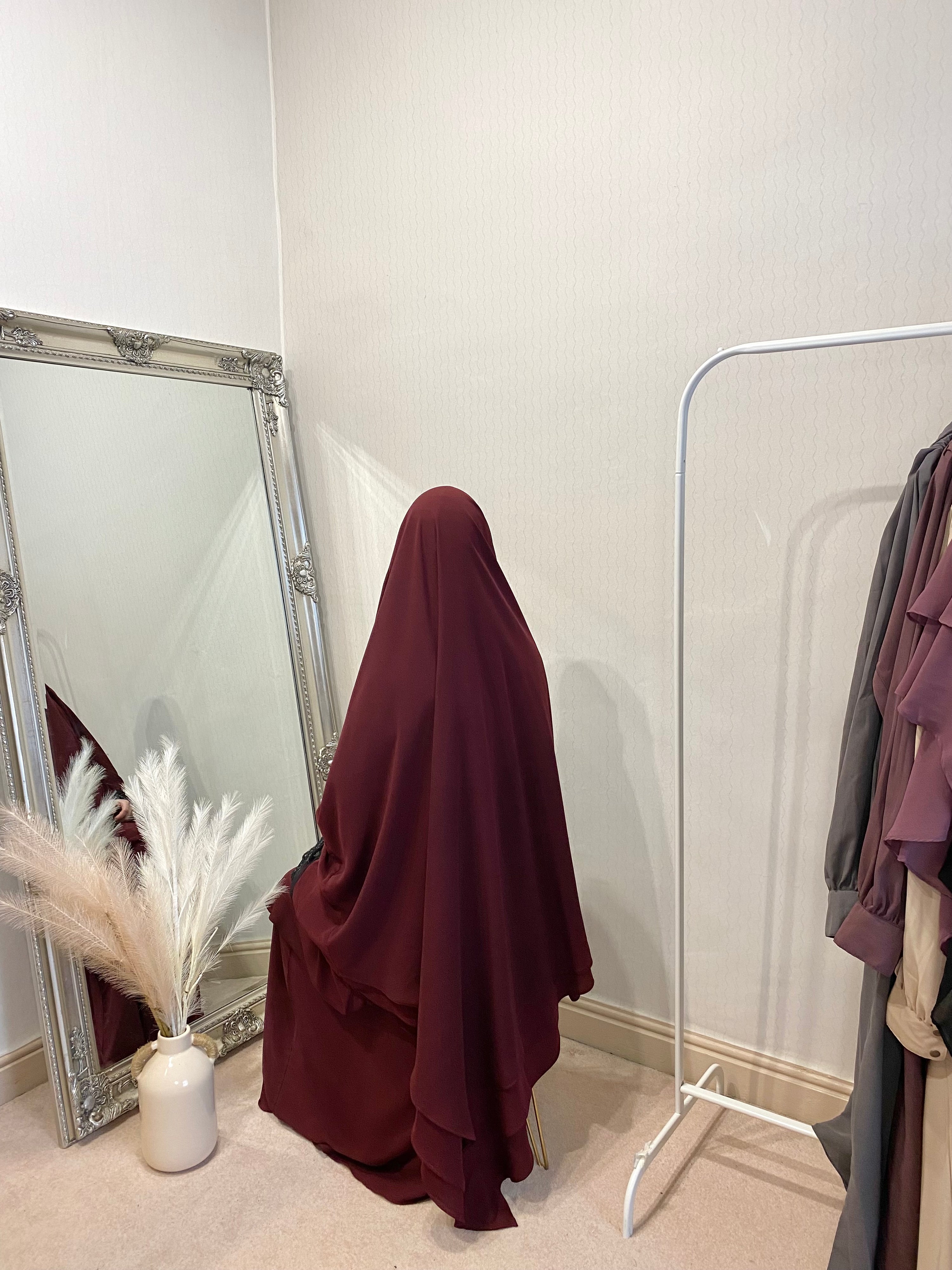 A person wearing a 3 layer jilbab in a room with a large mirror, reflecting the person and some clothing items. The jilbab is in solid color and the person is seated on a chair.