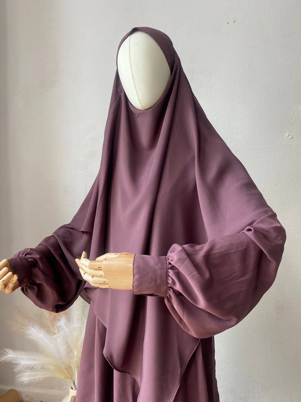 A woman wearing a purple abaya and khimar set with a tieback headband, standing indoors with her back towards the camera.