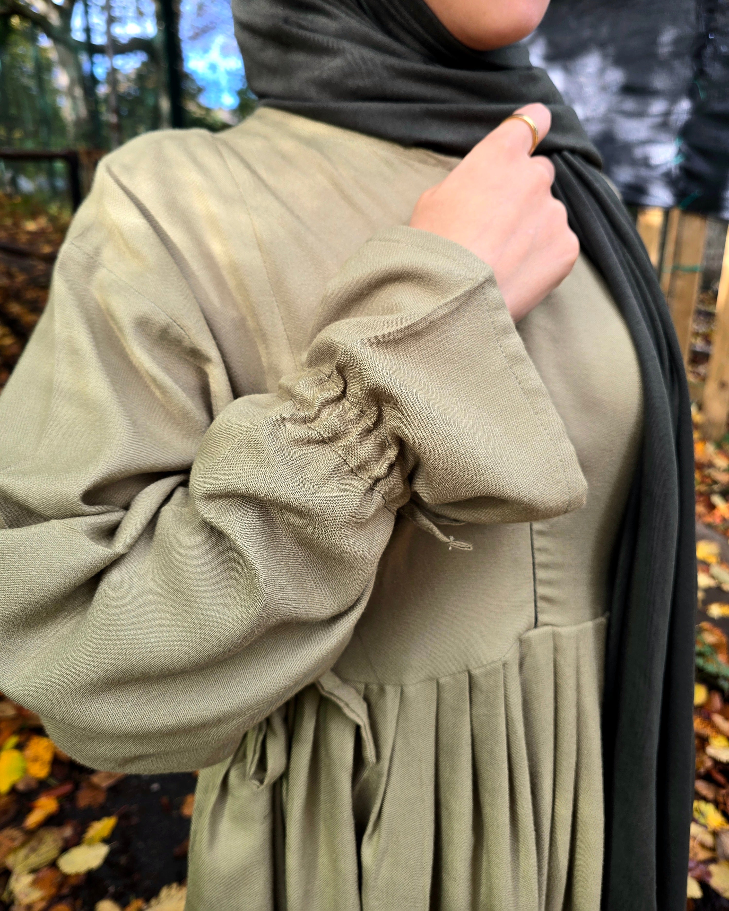 Person wearing a green dress with long sleeves and a dark hijab in an outdoor setting with trees and leaves.