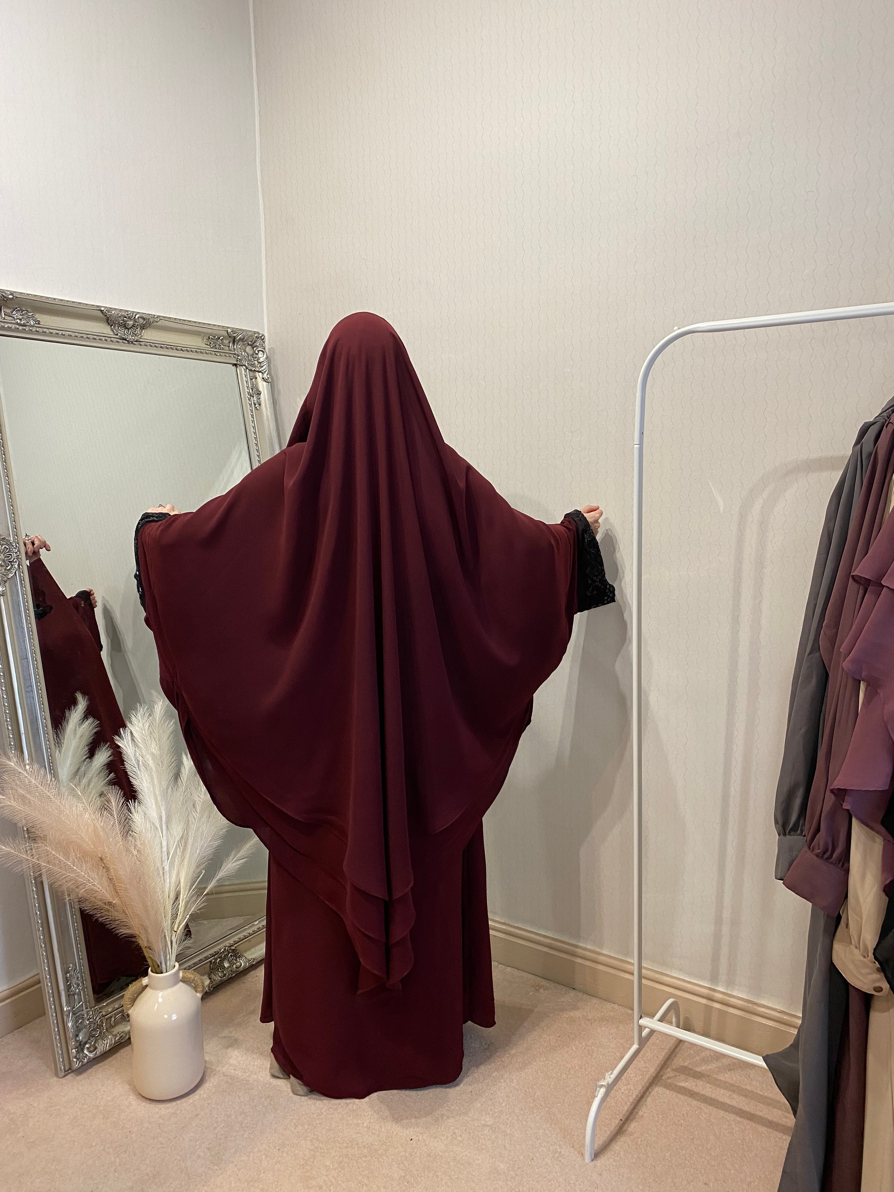 A person wearing a 3 layer jilbab in a room with a large mirror, reflecting the person and some clothing items. The jilbab is in solid color and the person is seated on a chair.