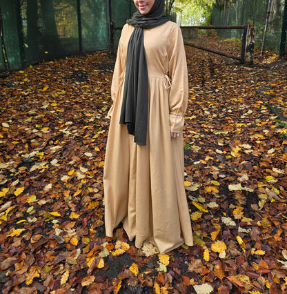 Person in a beige cotton abaya and dark green jersey headscarf standing on a path with autumn leaves