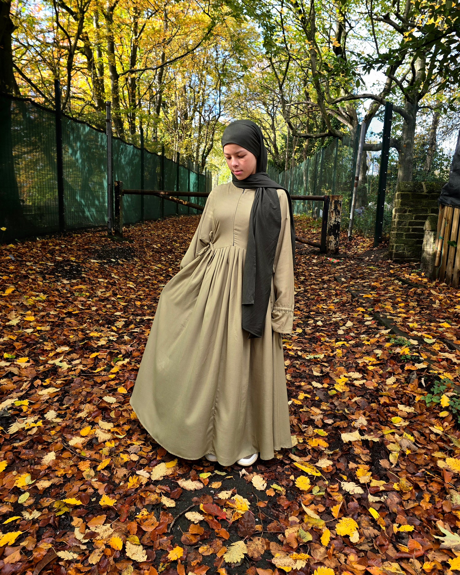 Person wearing a abaya  dress and black hijab standing on a path with autumn leaves and trees.