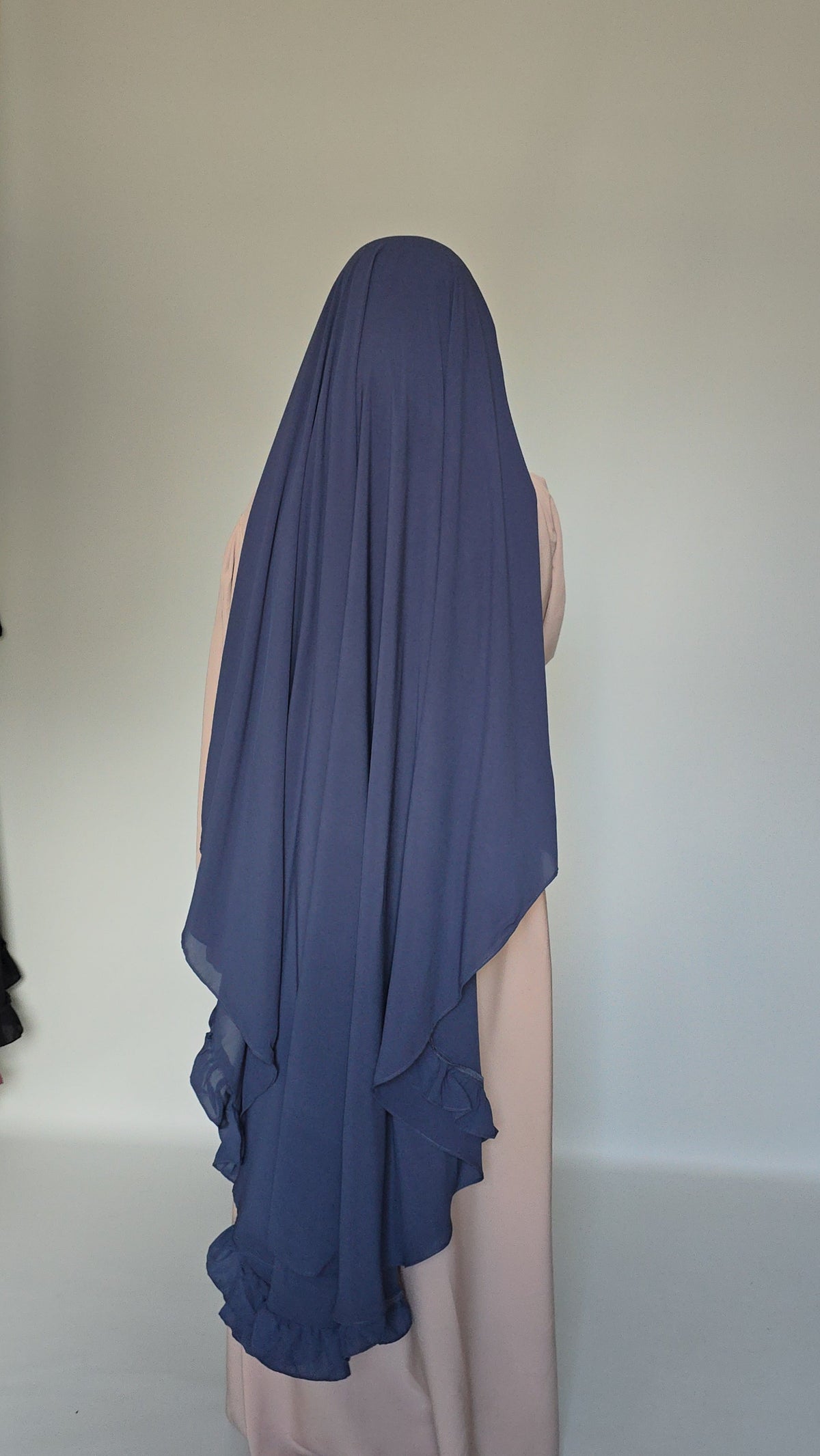 A navy blue chiffon hijab with layered ruffles, shown from the back on a mannequin wearing a light pink dress against a plain light gray background.
