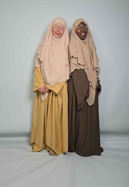 Two individuals wearing beige hijabs and long dresses on a plain background