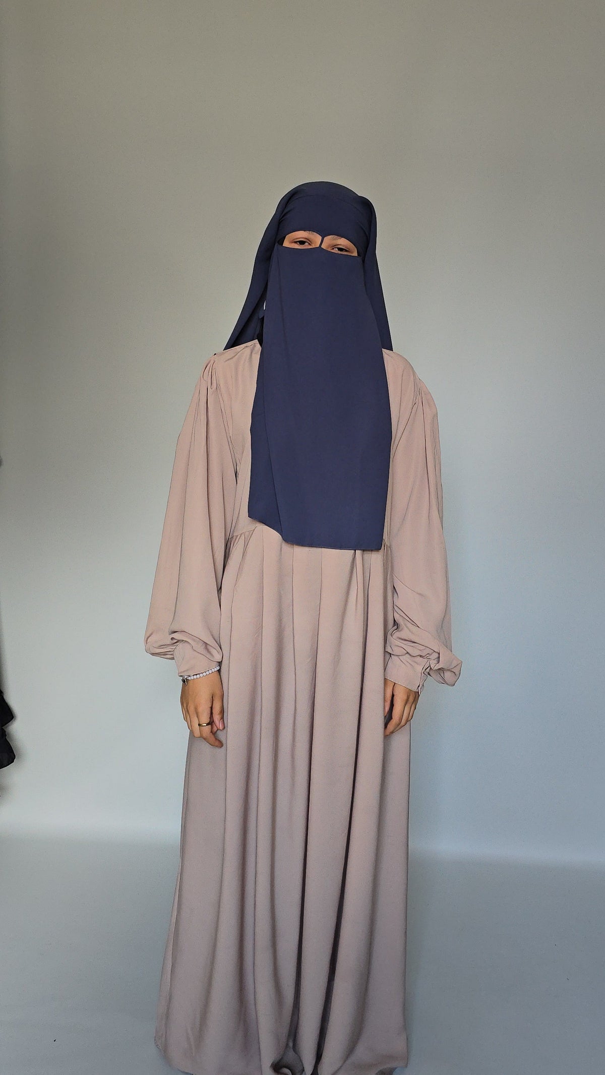 A woman wearing a long, flowing abaya dress in a muted cream with a matching navy blue niqab head covering, standing against a plain light gray studio background.