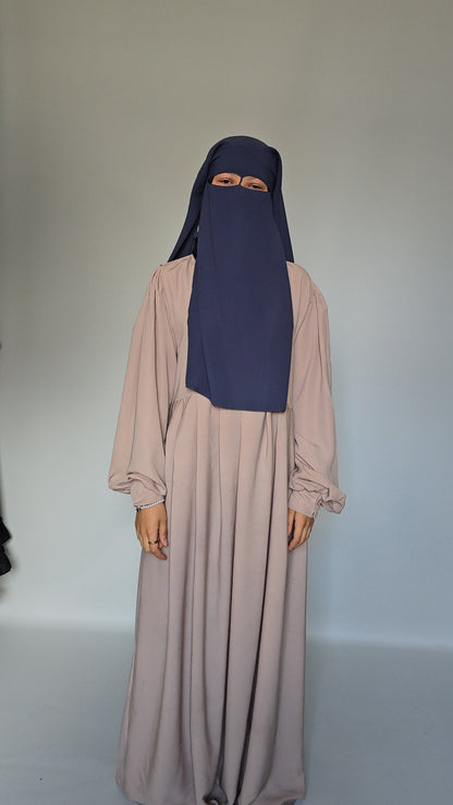 A woman wearing a long, flowing abaya dress in a muted cream with a matching navy blue niqab head covering, standing against a plain light gray studio background.