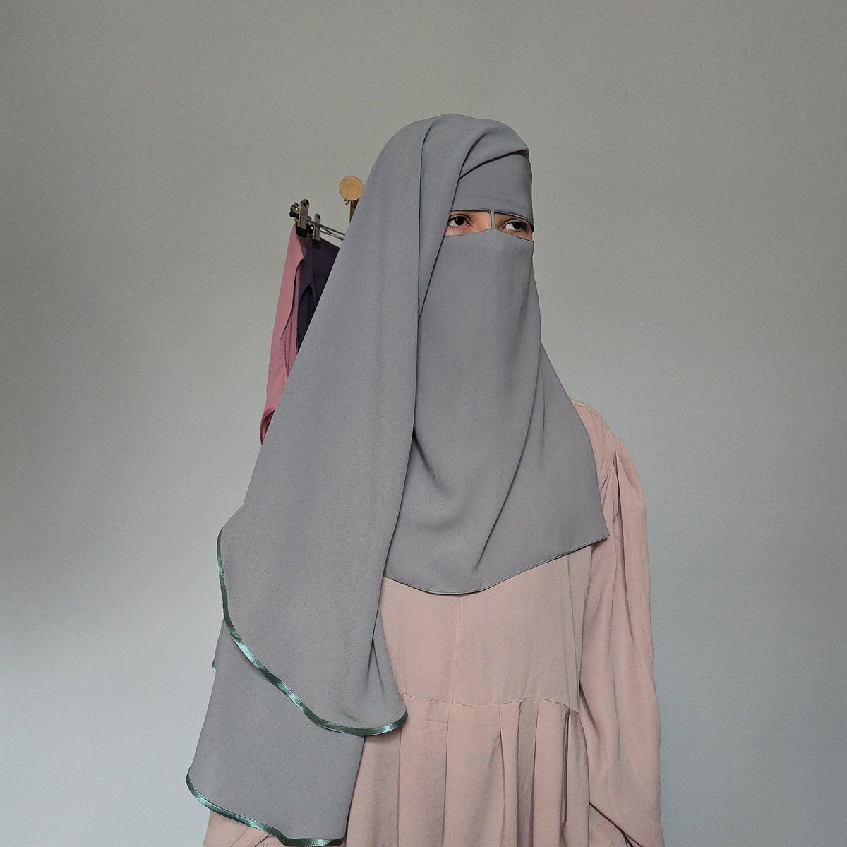 A person wearing a light mint full-face hijab and a long cream abaya, standing against a white studio backdrop.