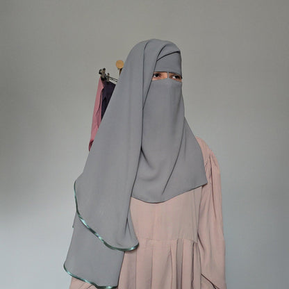A person wearing a light mint full-face hijab and a long cream abaya, standing against a white studio backdrop.