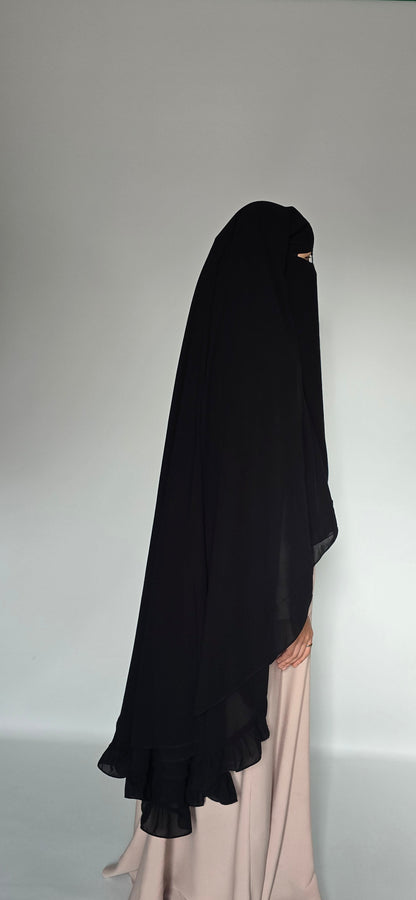 A woman wearing a sheer black full-body modest garment with a ruffled hem, shown in side profile against a plain light gray studio background.