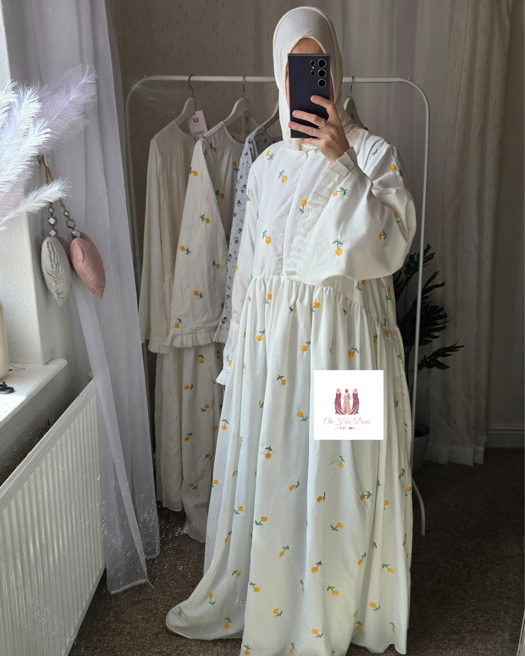 Person wearing a white dress with yellow  little buttercups patterns in a room with white curtains and clothing on hangers.