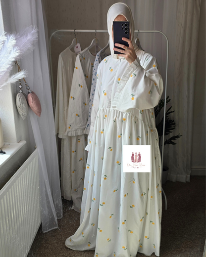 Person wearing a white dress with yellow  little buttercups patterns in a room with white curtains and clothing on hangers.