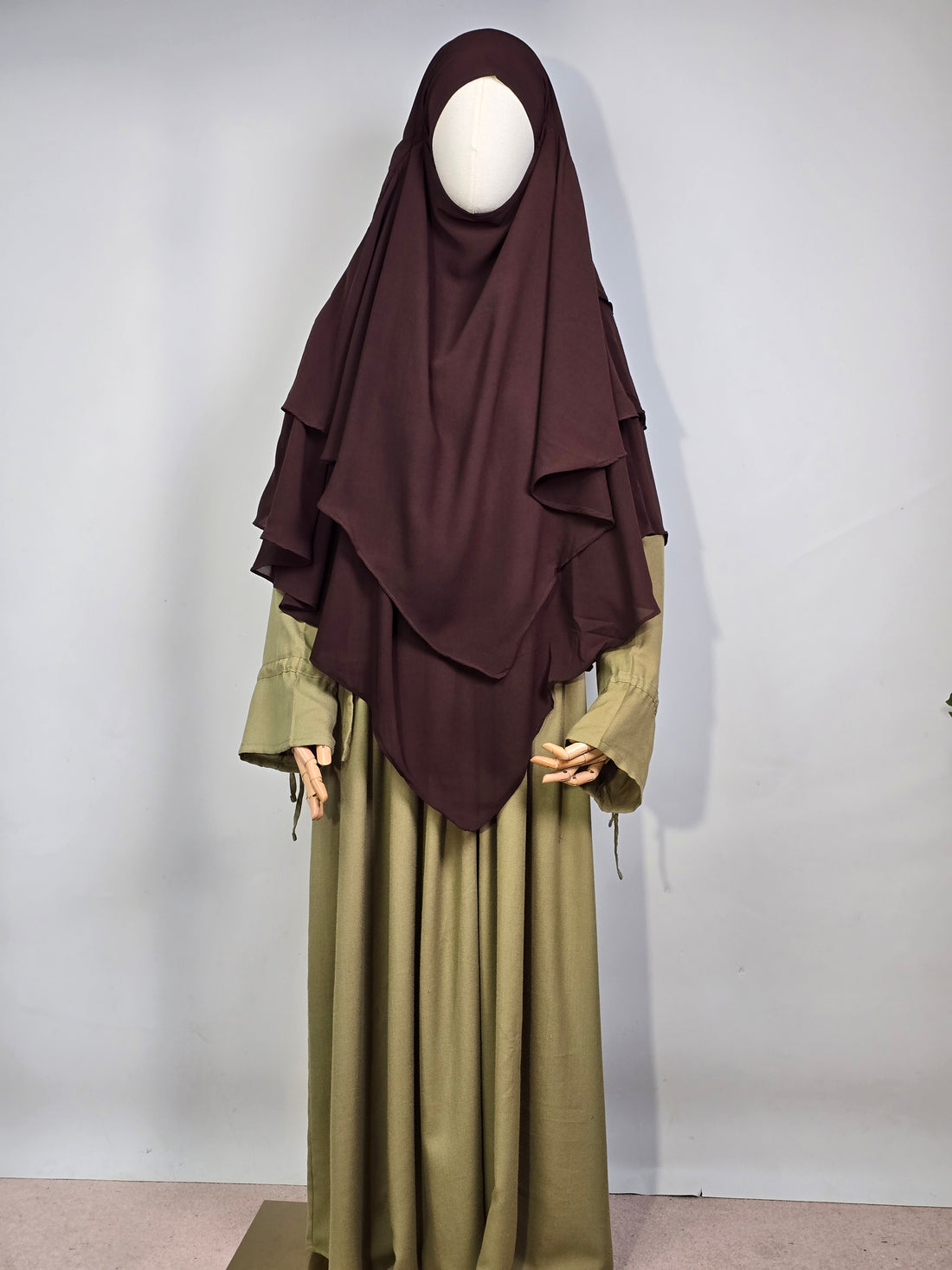 mannequin wearing a dark brown layered chiffon hijab khimar with a green dress on a white background 