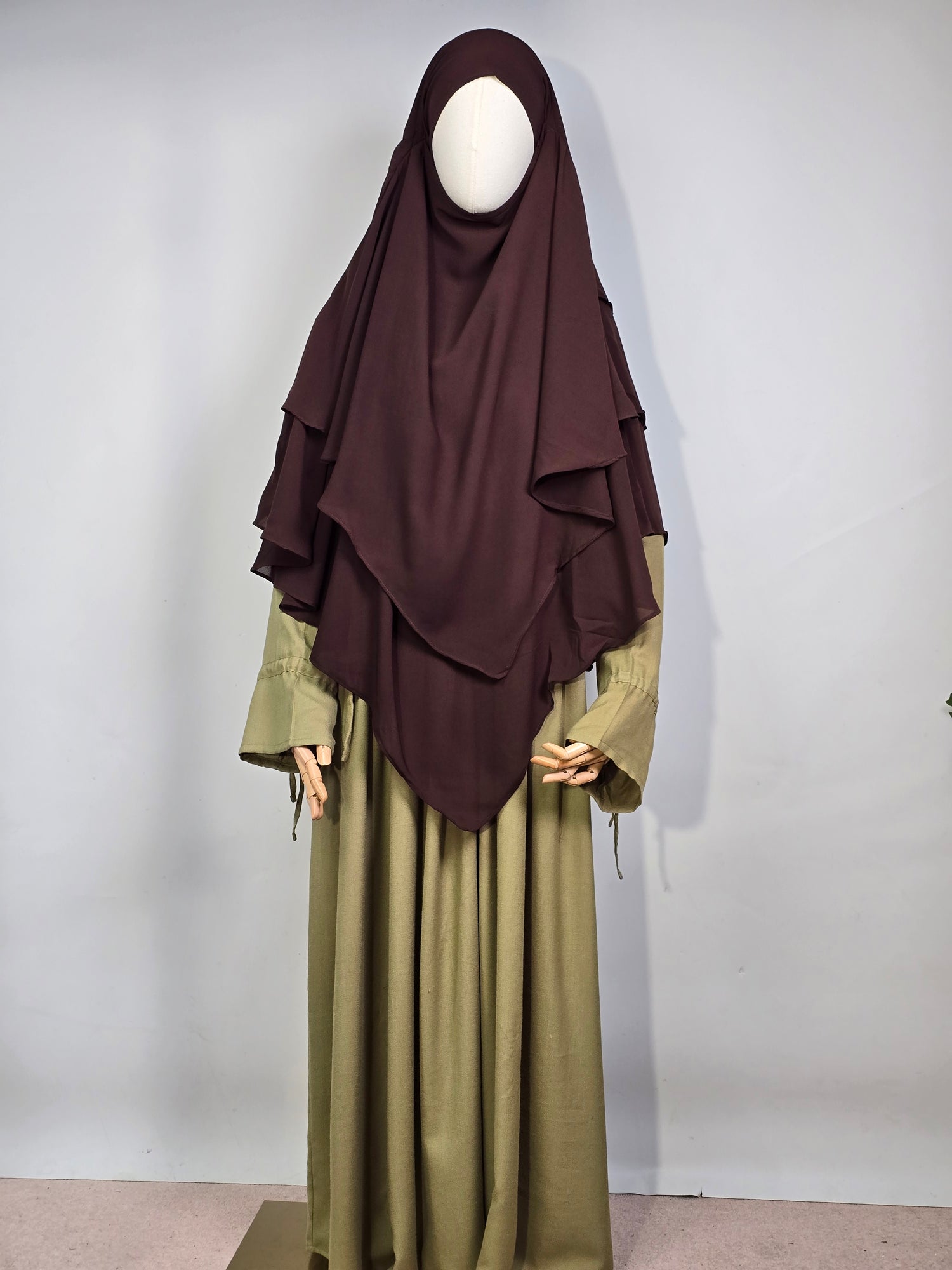 mannequin wearing a dark brown layered chiffon hijab khimar with a green dress on a white background 
