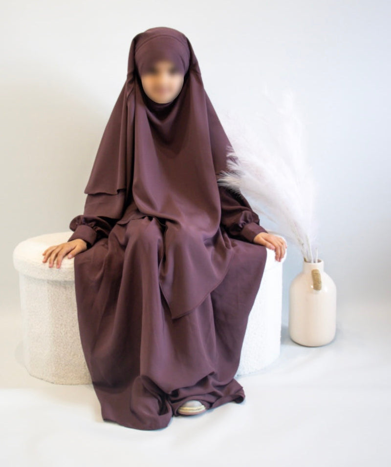 Person wearing a dusky rose  abaya &amp; sitting on a white stool against background 