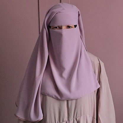 A person wearing a light purple niqab face veil over a beige dress, standing against a matching dusty pink background.