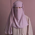 A person wearing a light purple niqab face veil over a beige dress, standing against a matching dusty pink background.