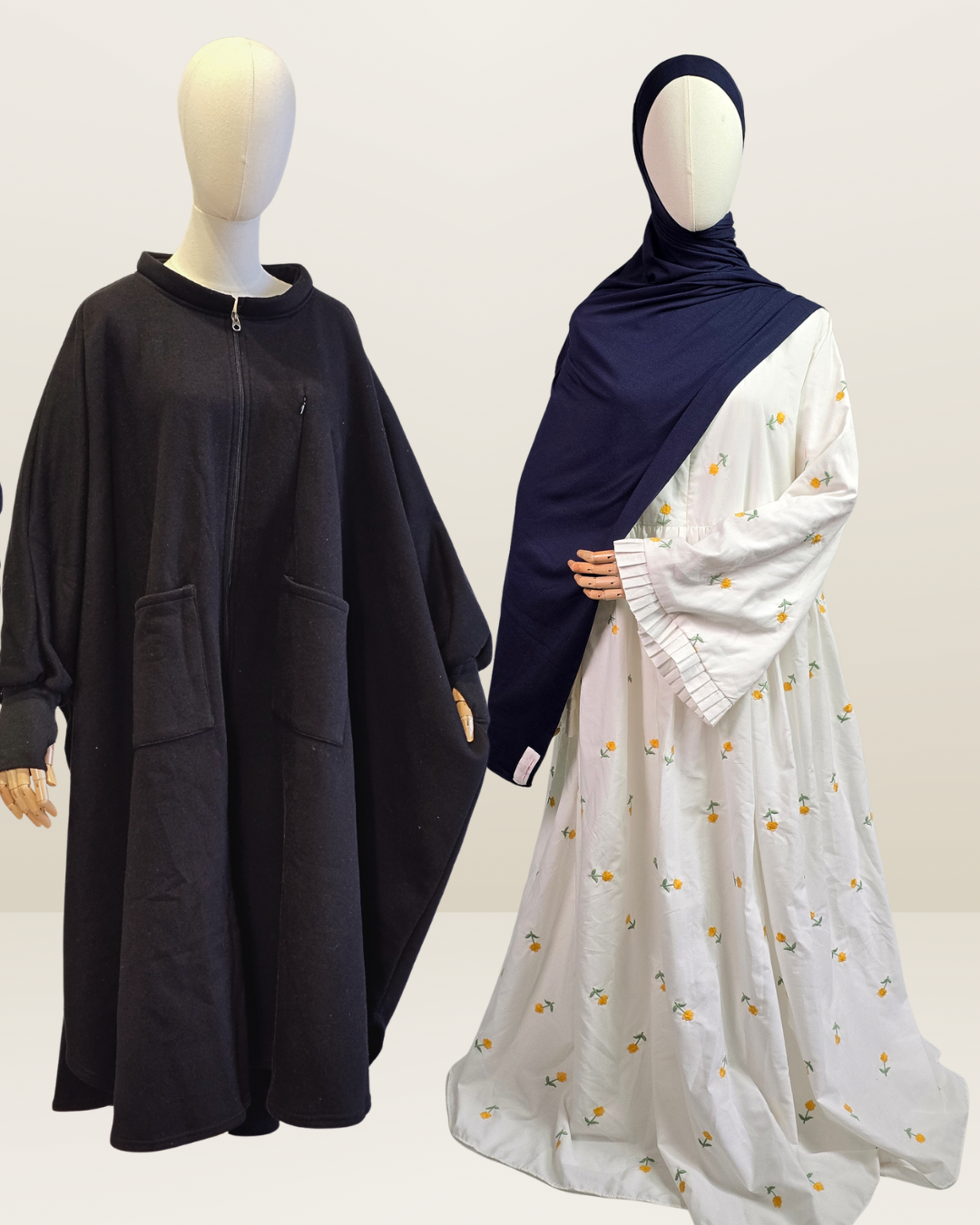 Two mannequins display modest clothing against a plain light background. The left mannequin wears a loose black abaya with a front zipper and large patch pockets.