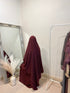 A person wearing a 3 layer jilbab in a room with a large mirror, reflecting the person and some clothing items. The jilbab is in solid color and the person is seated on a chair.