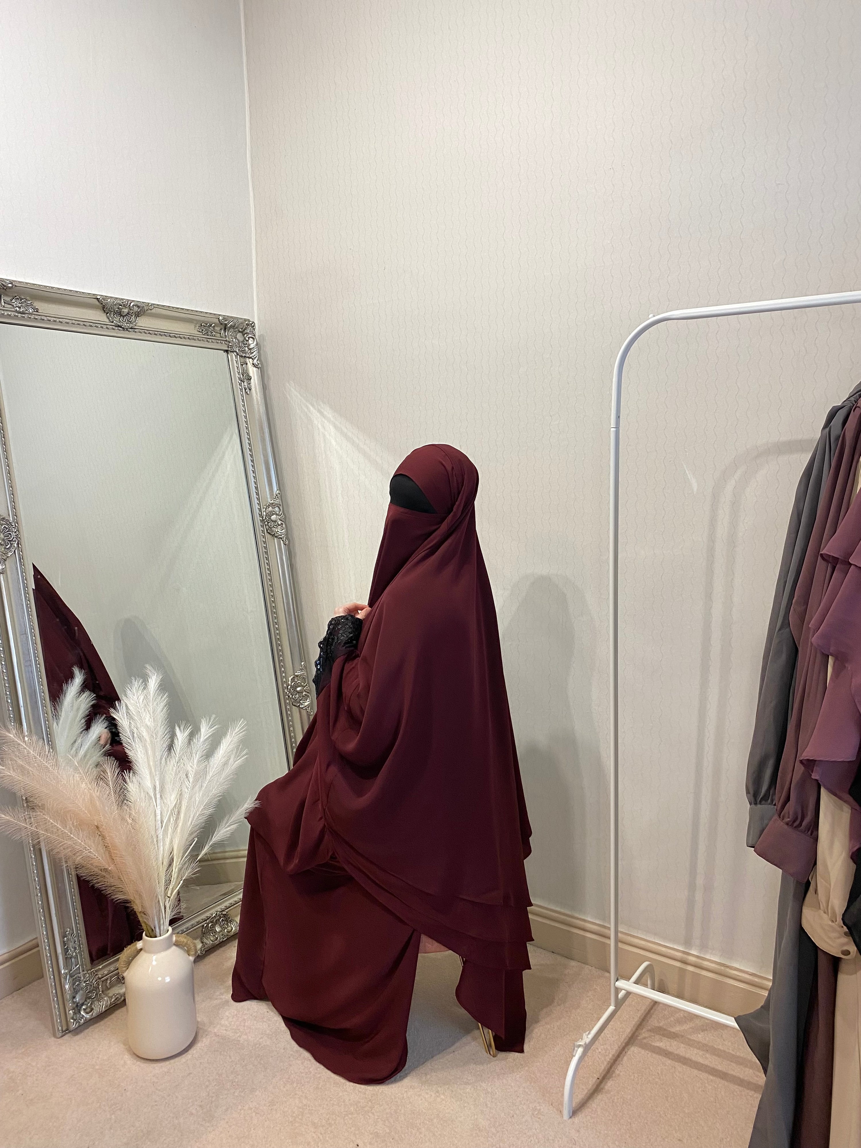 A person wearing a 3 layer jilbab in a room with a large mirror, reflecting the person and some clothing items. The jilbab is in solid color and the person is seated on a chair.