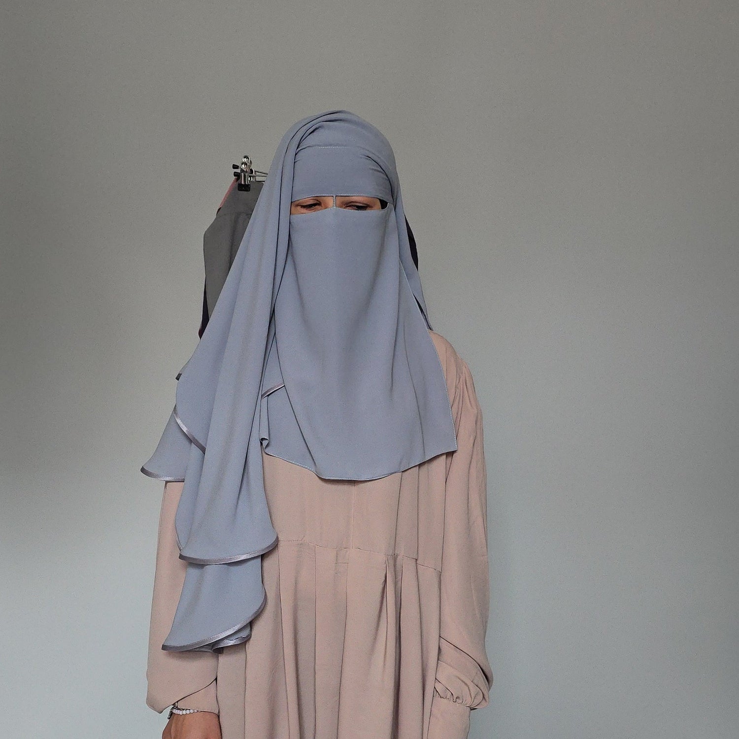 A person wearing a light blue-gray hijab and a long, flowing dusty pink abaya, standing against a light gray studio backdrop.
