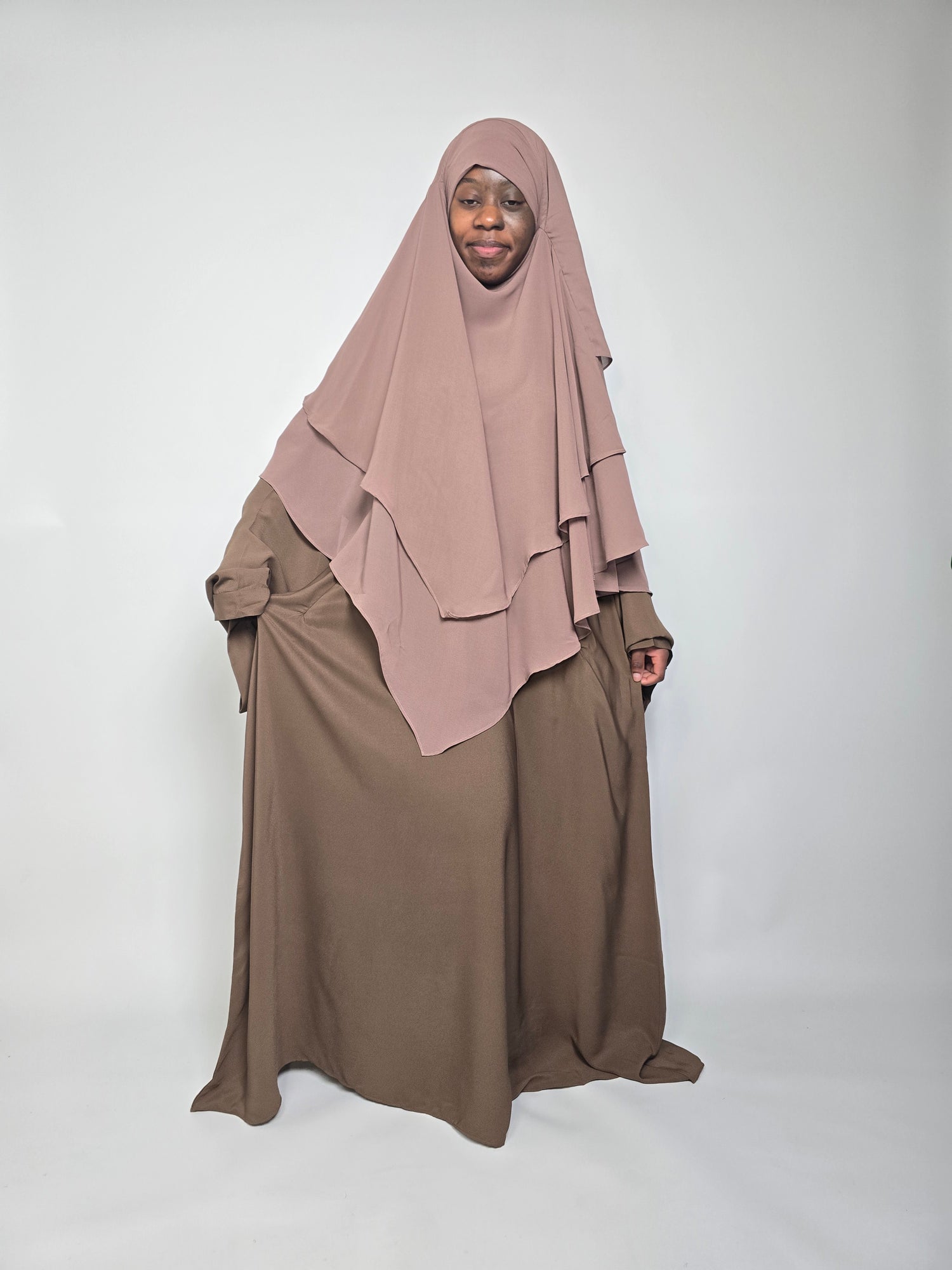 Woman in brown abaya with taupe chiffon khimar hijab stood on a white background 