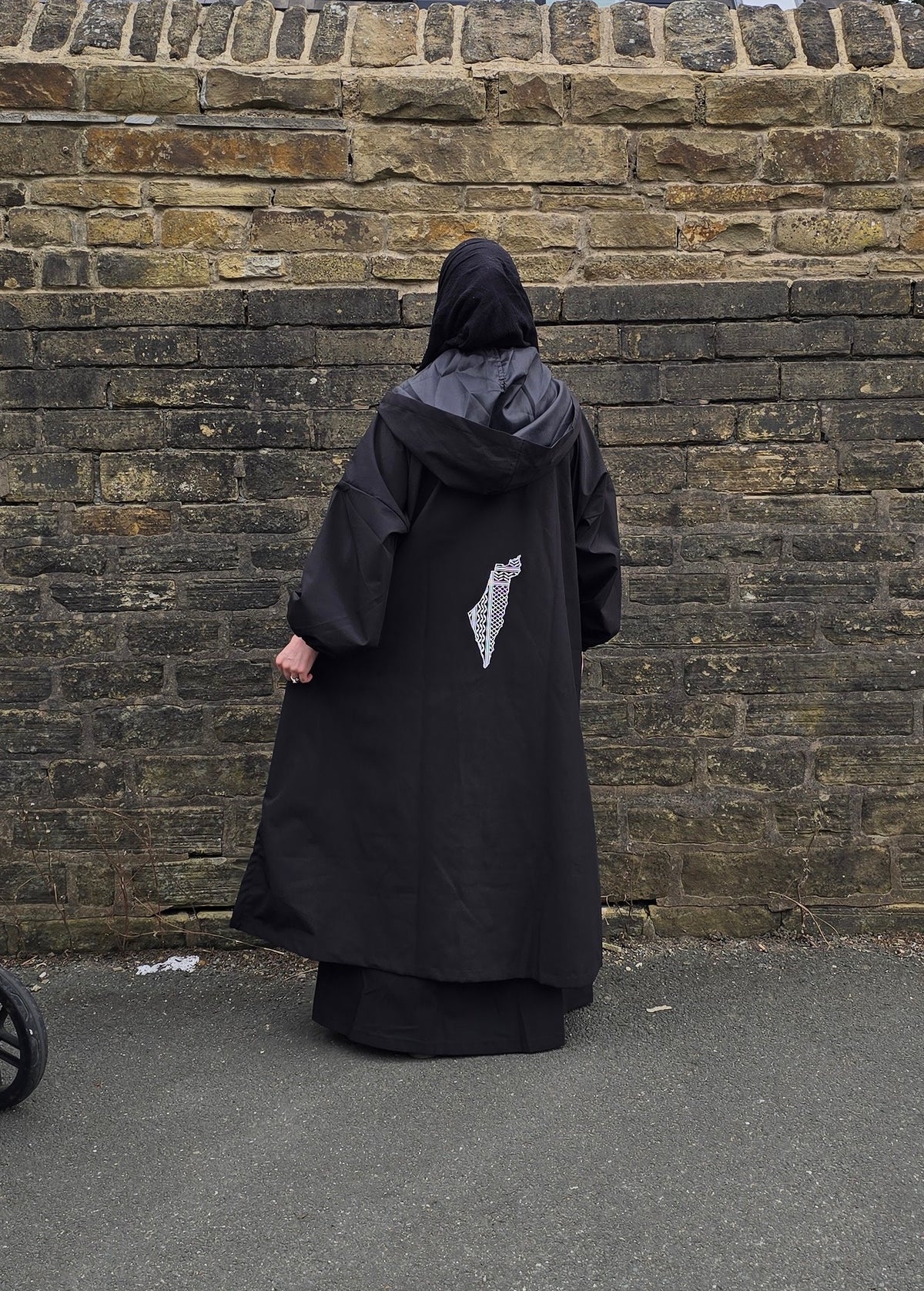 A person viewed from behind wearing a long black hooded robe or cloak with a small white graphic on the back, standing in front of a weathered stone wall on pavement.