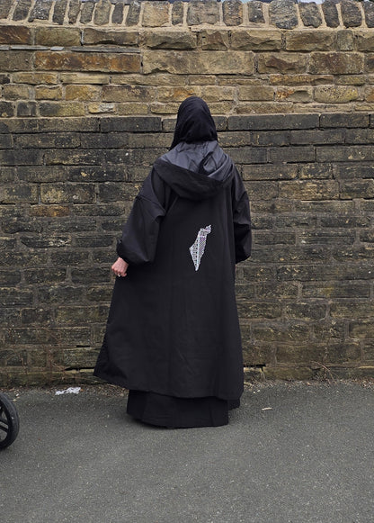A person viewed from behind wearing a long black hooded robe or cloak with a small white graphic on the back, standing in front of a weathered stone wall on pavement.