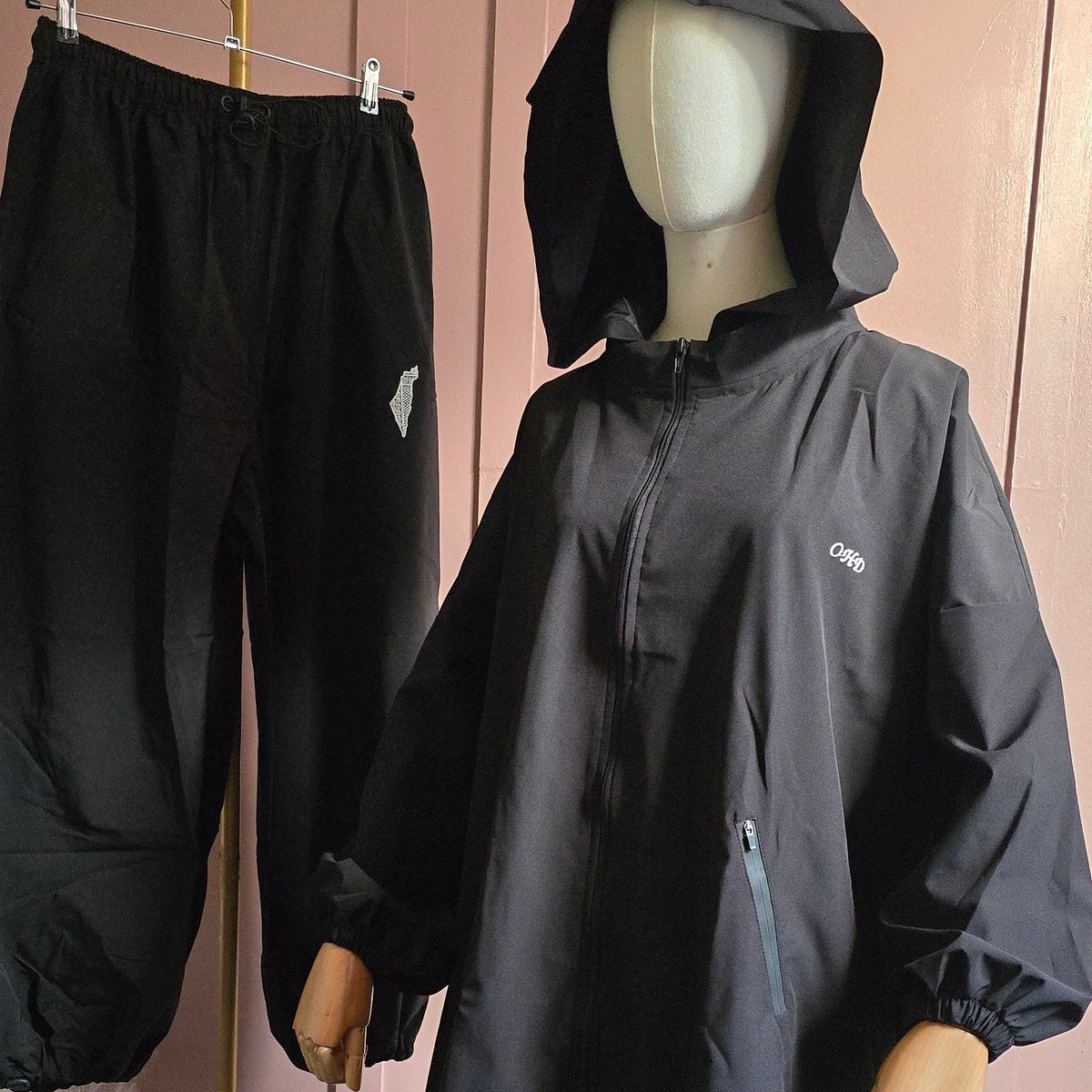 A black hooded rain poncho displayed on a mannequin, with matching black pants hanging on a stand to the left, set against a pink paneled wall.