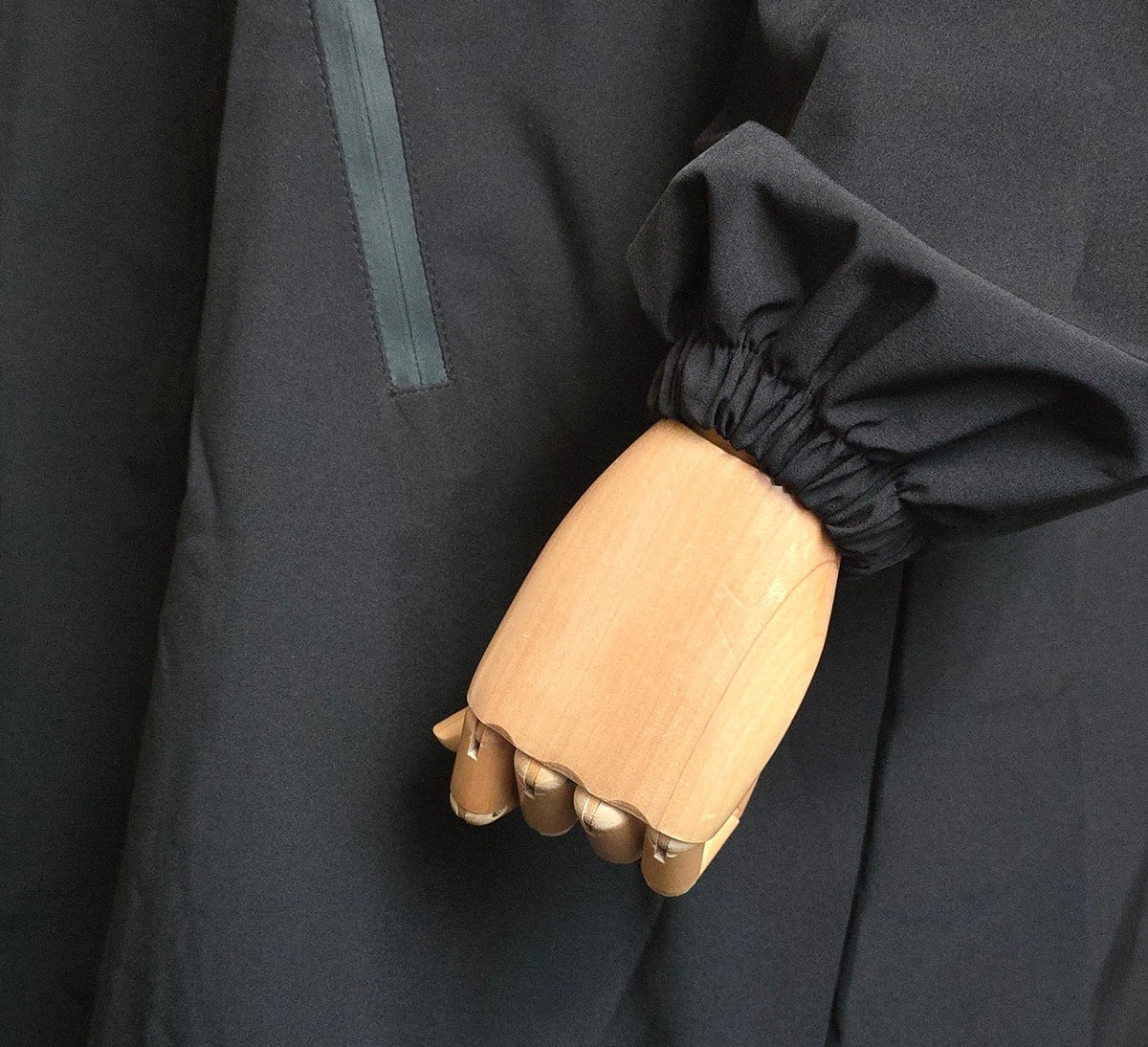 A close-up of a black hooded jacket or coat with a long sleeve and elasticized cuff, shown on a mannequin with a wooden hand. A vertical reflective-looking zipper detail is visible on the upper chest.