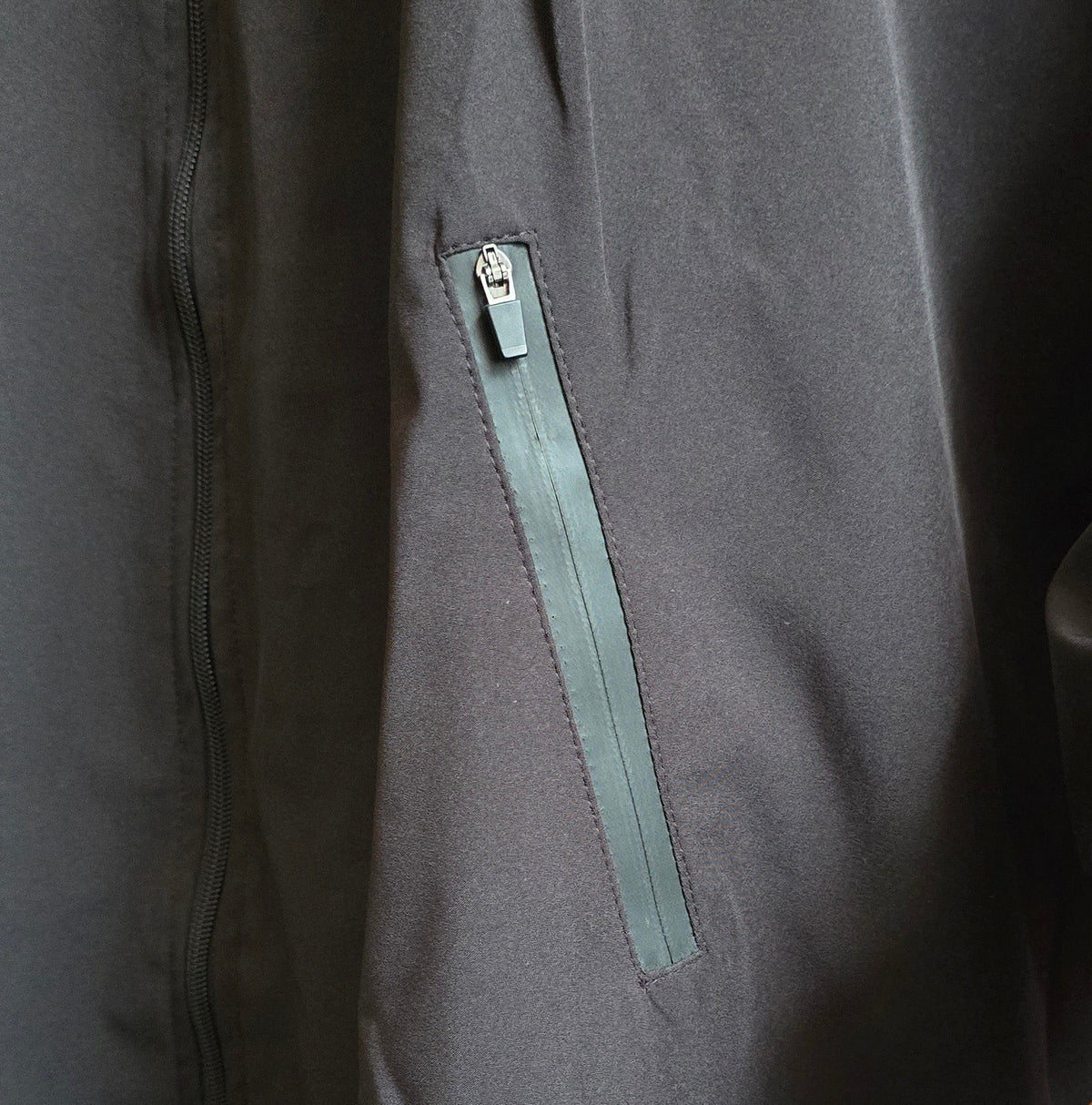 Close-up of a dark gray jacket or coat with a vertical zippered pocket featuring a muted green zipper pull, shown on a mannequin with a wooden hand visible at the lower right.