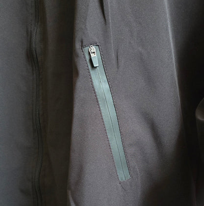 Close-up of a dark gray jacket or coat with a vertical zippered pocket featuring a muted green zipper pull, shown on a mannequin with a wooden hand visible at the lower right.