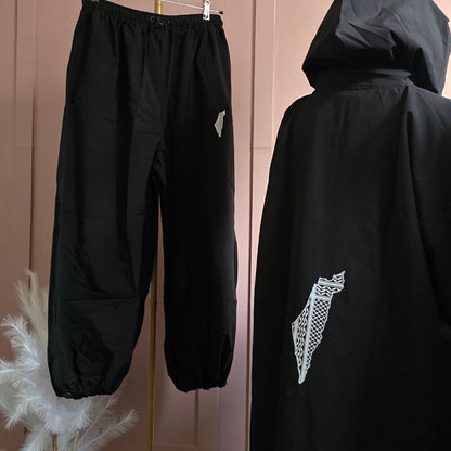 Black jogger pants hanging on a gold stand against a pink arched wall, next to a black hooded jacket with a white graphic print.
