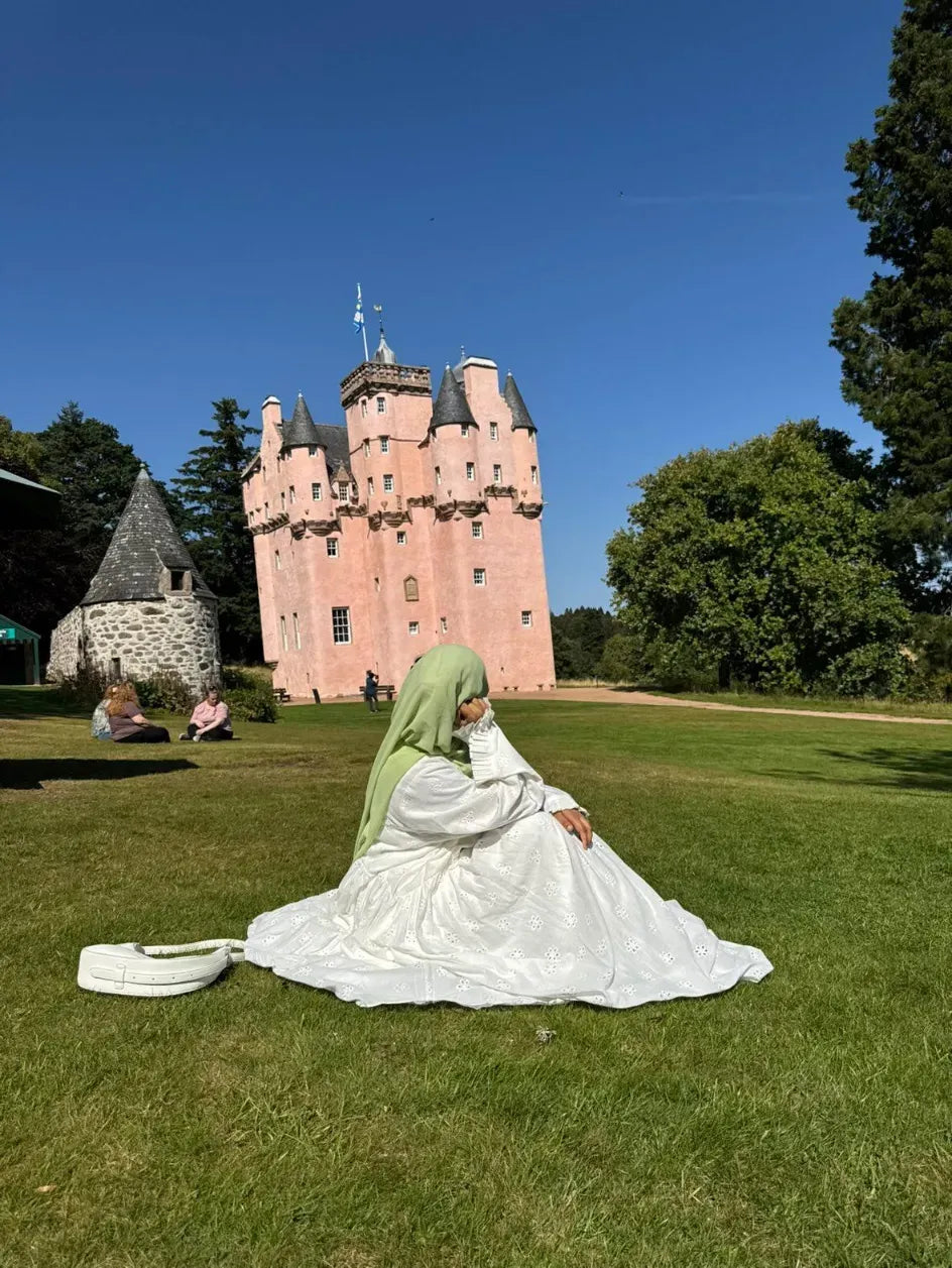 Women sat on grass in aeyelet cotto. About infront  of a pink castle 
