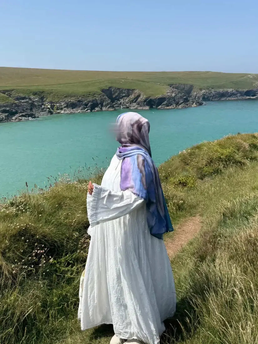 A person wearing a long white dress and a purple, blue, and white patterned hijab stands on a grassy cliffside overlooking a turquoise sea and green hills under a clear blue sky.
