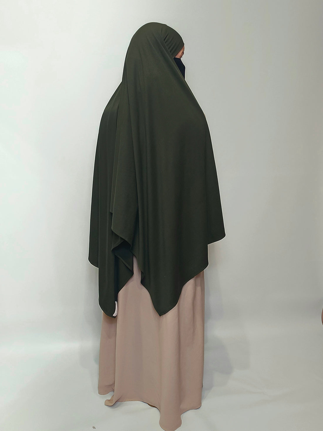 Person wearing a dark green jersey hijab over a beige dress on a white background