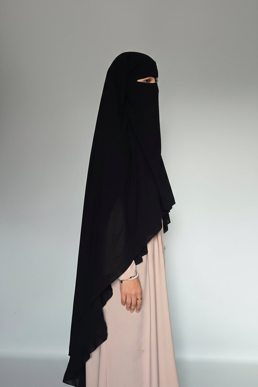 A woman wearing a black full-face burkini or niqab with a small eye slit, paired with a light blush pink abaya, standing against a plain light gray studio background.