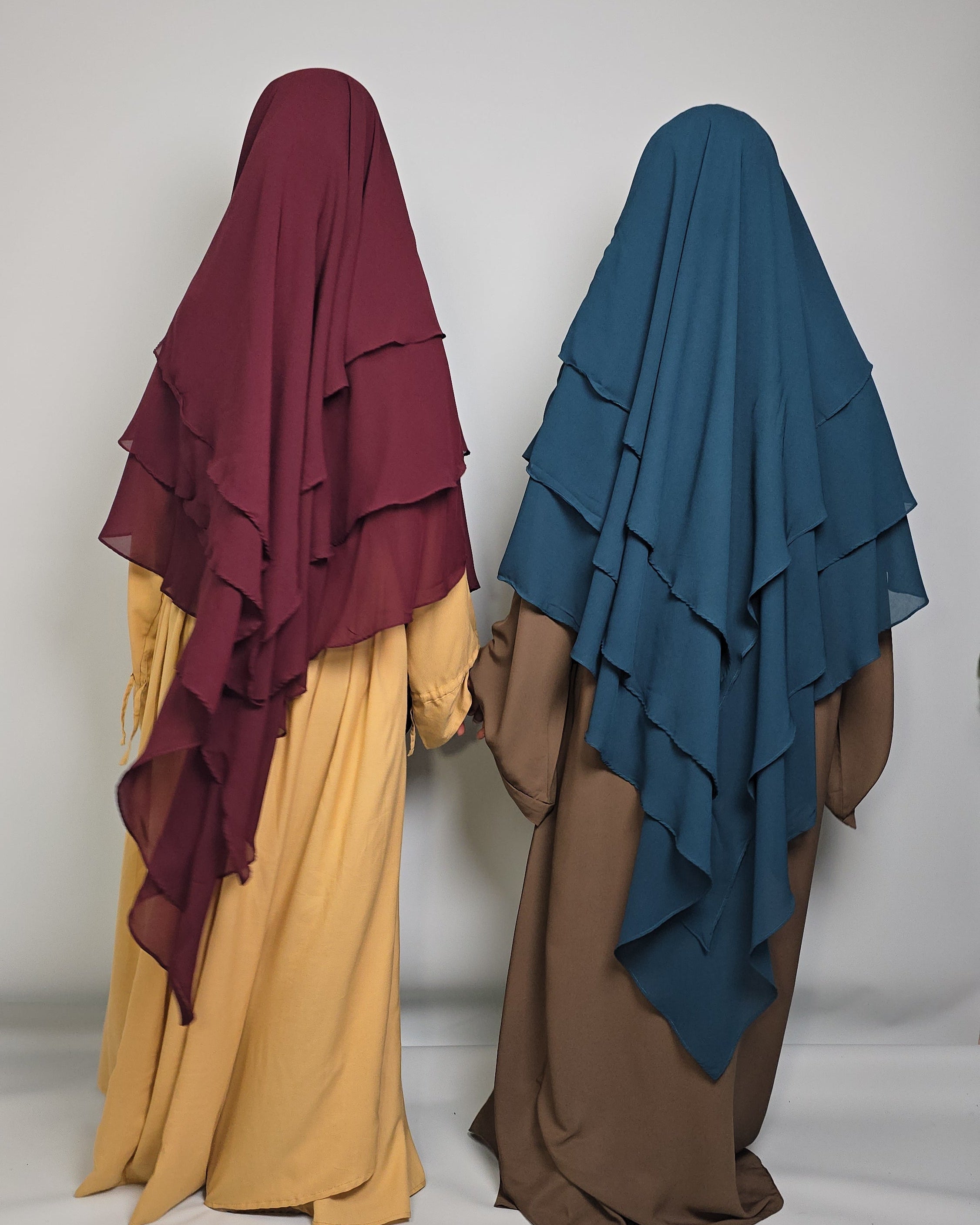 Two women wearing chiffon layer khimars in burgundy and petrol teal blue matched with abaya n a white background 