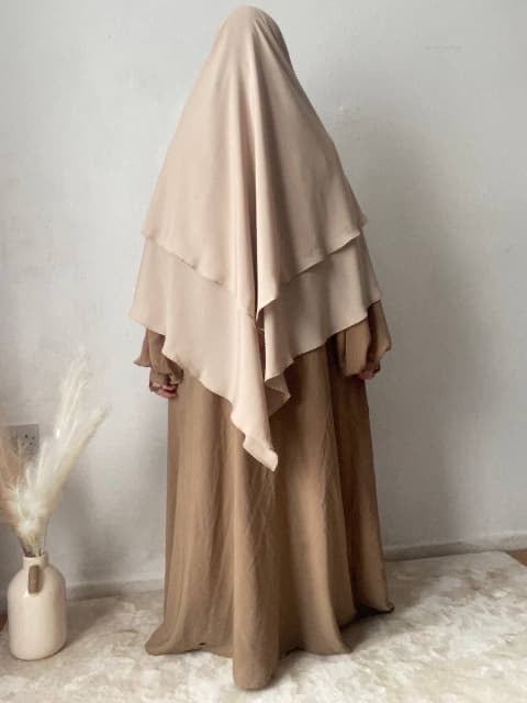 A person wearing a musk cream colored two-layer triangle khimar with niqab ties, standing indoors with a decorative item beside them.