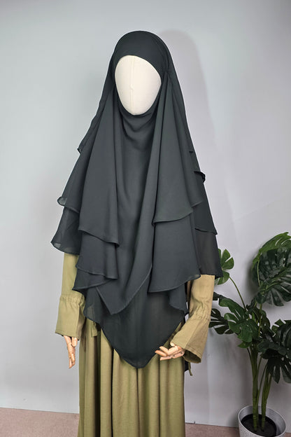 Person wearing a dark green khimar hijab with a sage green abaya with green plants against a plain background