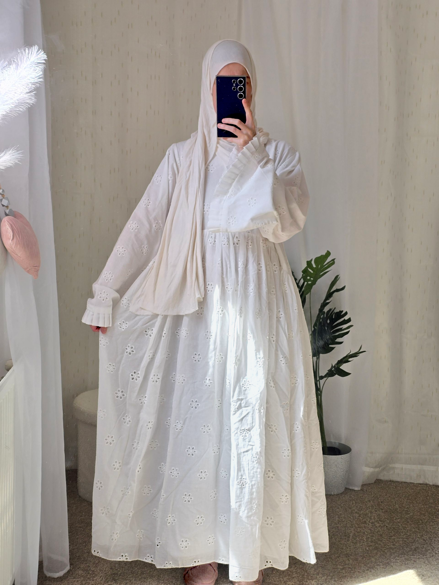 Person wearing a white cotton fully lined abaya dress with floral patterns in a room with beige curtains and a plant