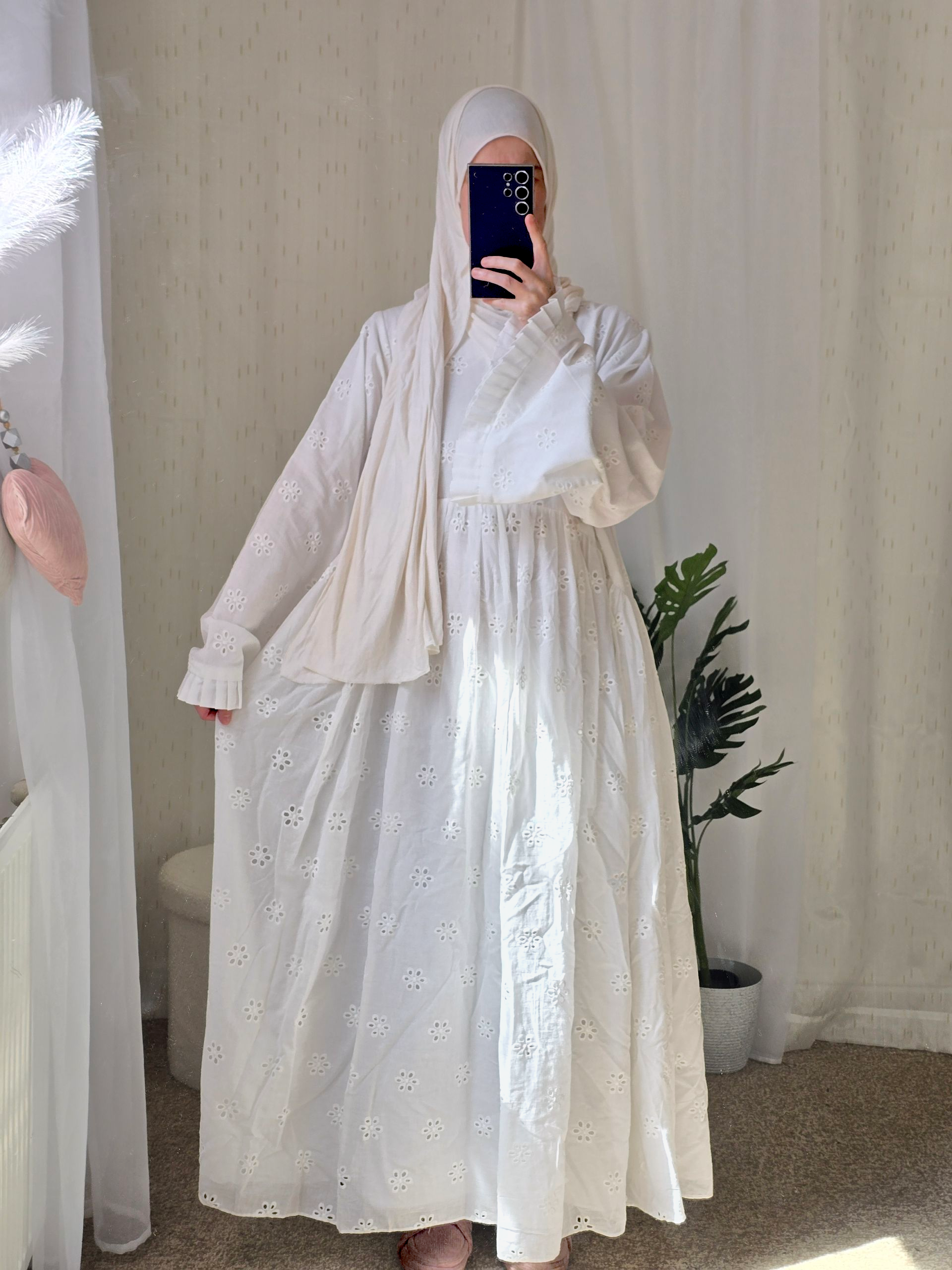 Person wearing a white cotton fully lined abaya dress with floral patterns in a room with beige curtains and a plant