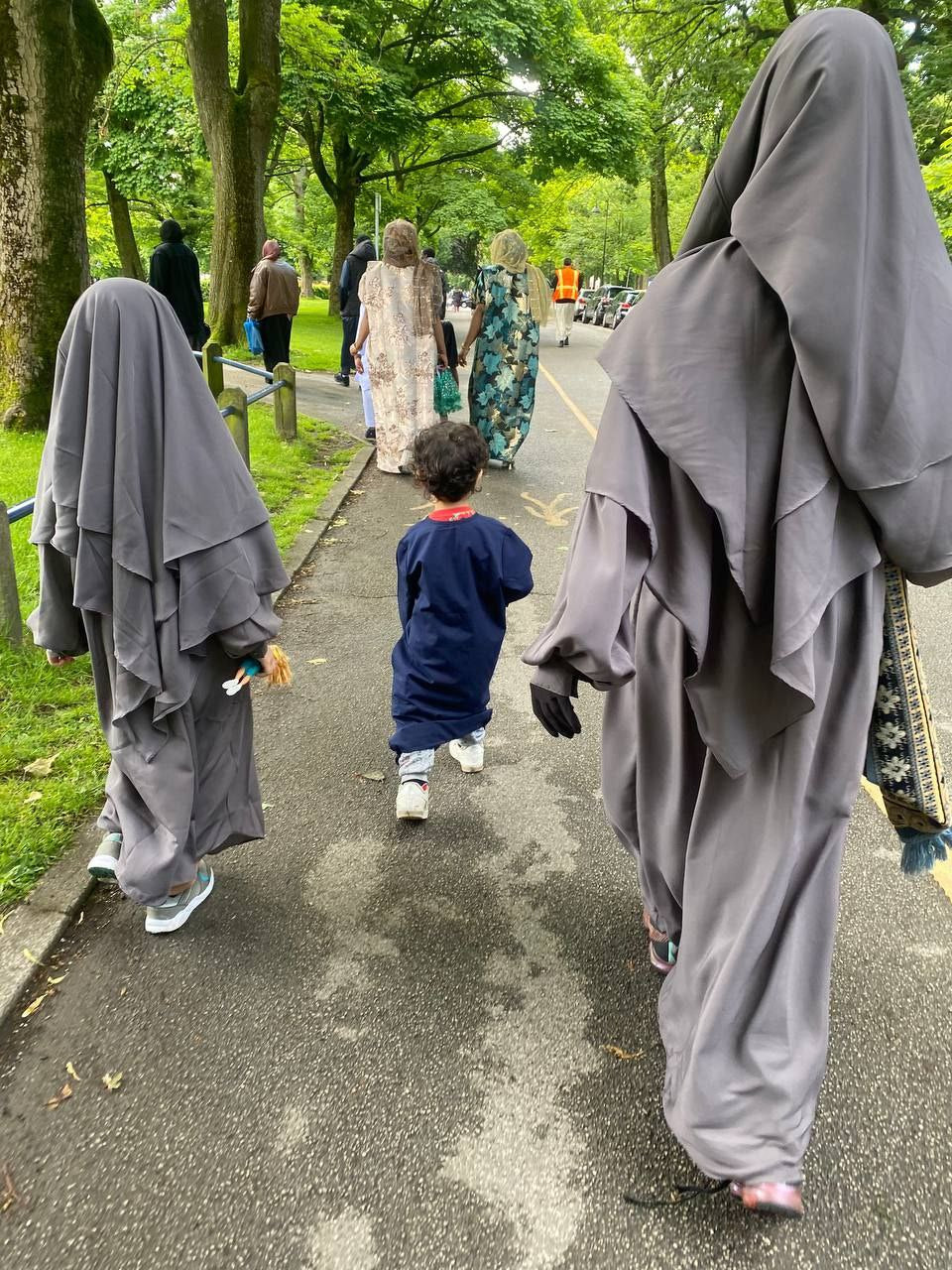 one girl and one women wearing grey, layered khimars with attached Russian abayas, walking on a pathway in a park.